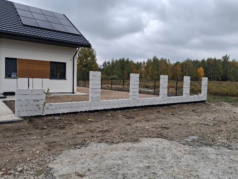 Wznoszona podmurówka ogrodzenia z szarych bloczków betonowych na tle domu z panelami słonecznymi i jesiennego krajobrazu. Widoczny fragment metalowego ogrodzenia.