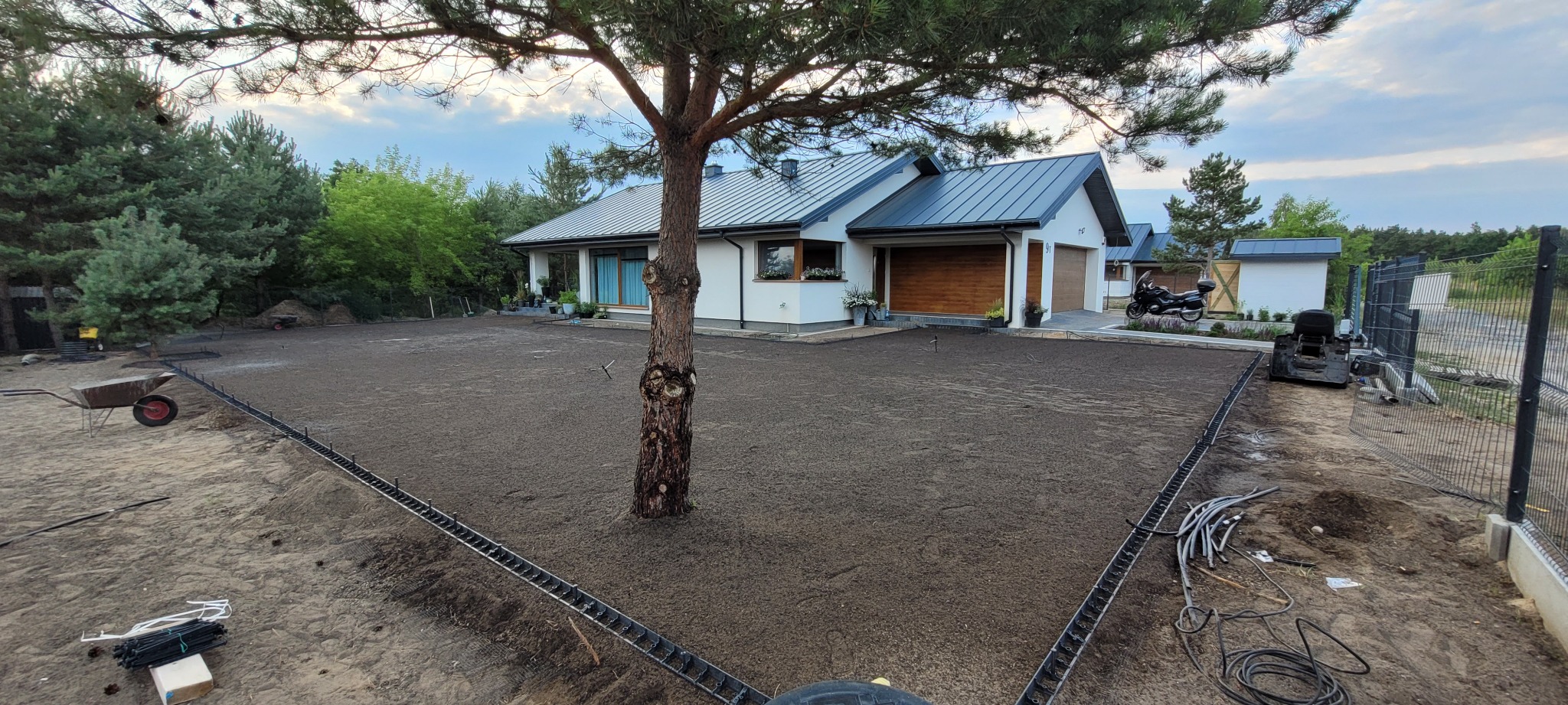 Przygotowany teren pod trawnik z systemem nawadniania, widok na dom z garażem i motocyklem. W tle zieleń drzew i krzewów, częściowo widoczne ogrodzenie.