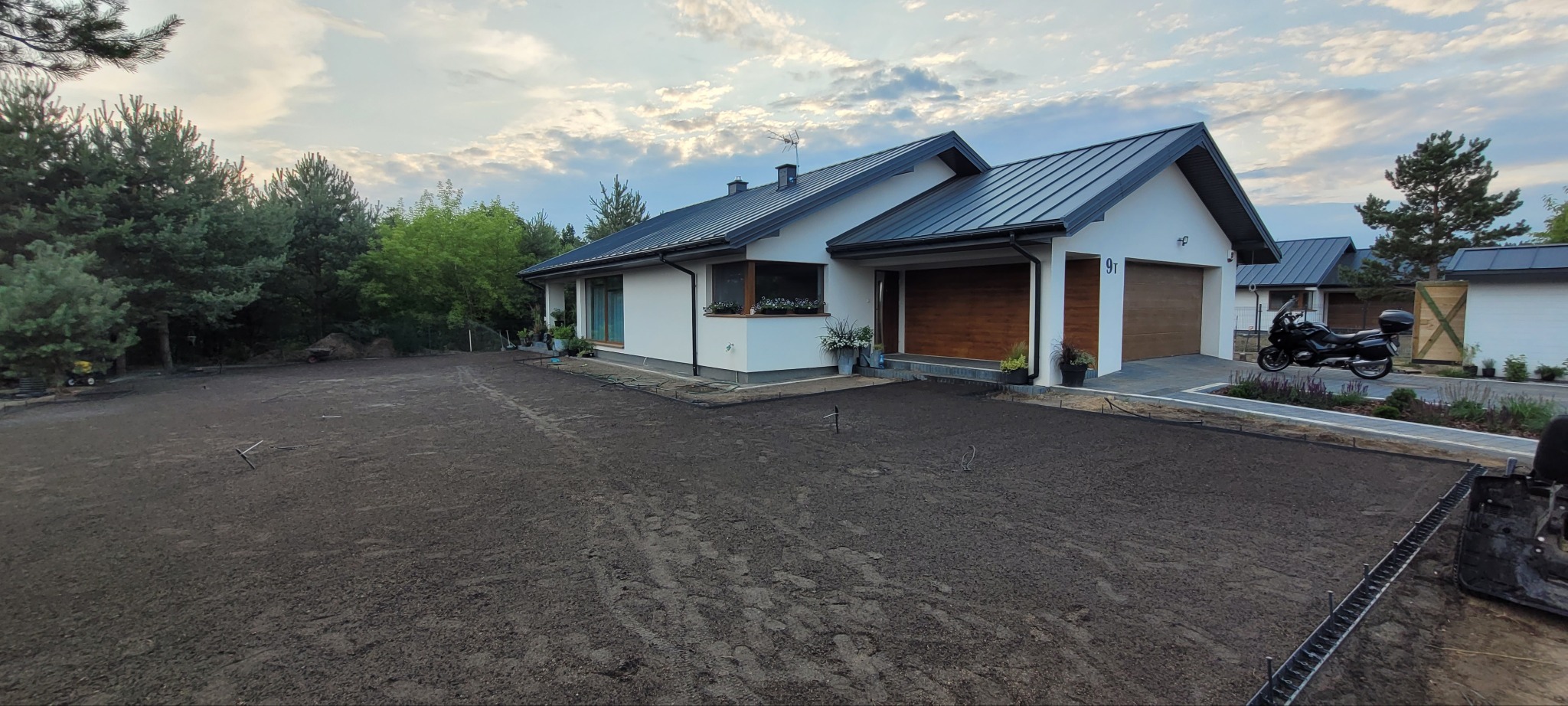 Przygotowany teren pod trawnik przed nowoczesnym domem z garażem i motocyklem. Widoczne ślady opon na ziemi i narzędzia ogrodnicze. Etap prac ogrodniczych.