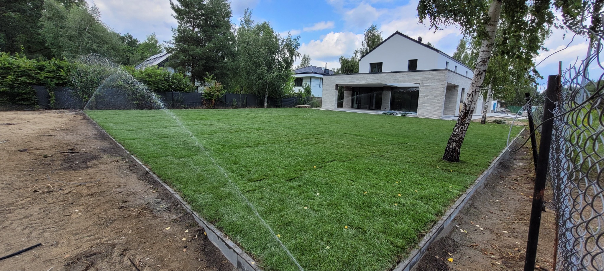 Nawadnianie trawnika z rolki w ogrodzie nowoczesnego domu. Widoczne zraszacze i świeżo ułożona trawa. Granica trawnika wykończona obrzeżem.