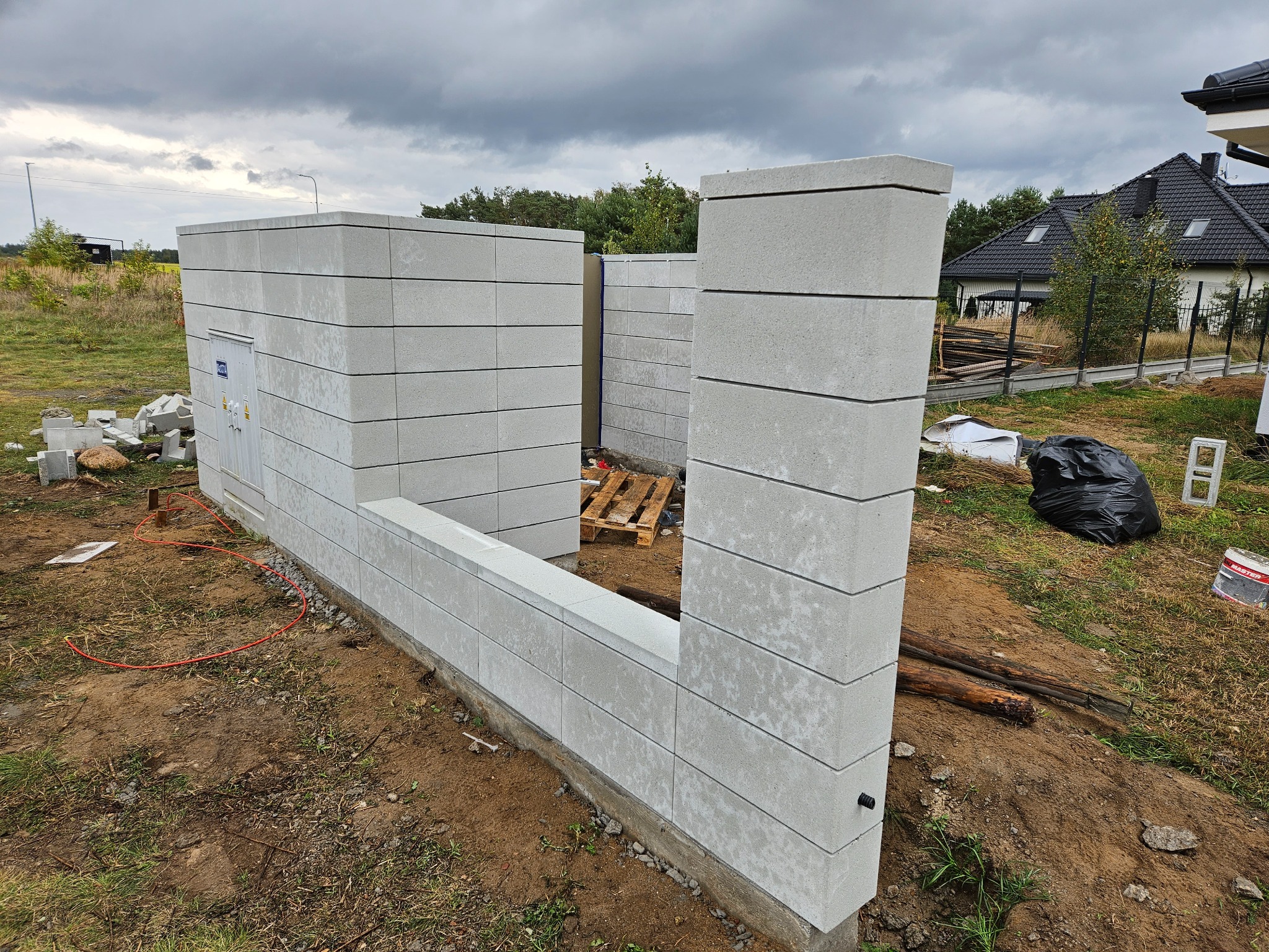 Wznoszona konstrukcja ogrodzenia z szarego bloczka betonowego, widoczne słupki i fragment muru. Na ziemi leżą narzędzia i materiały budowlane. Pochmurne niebo w tle.