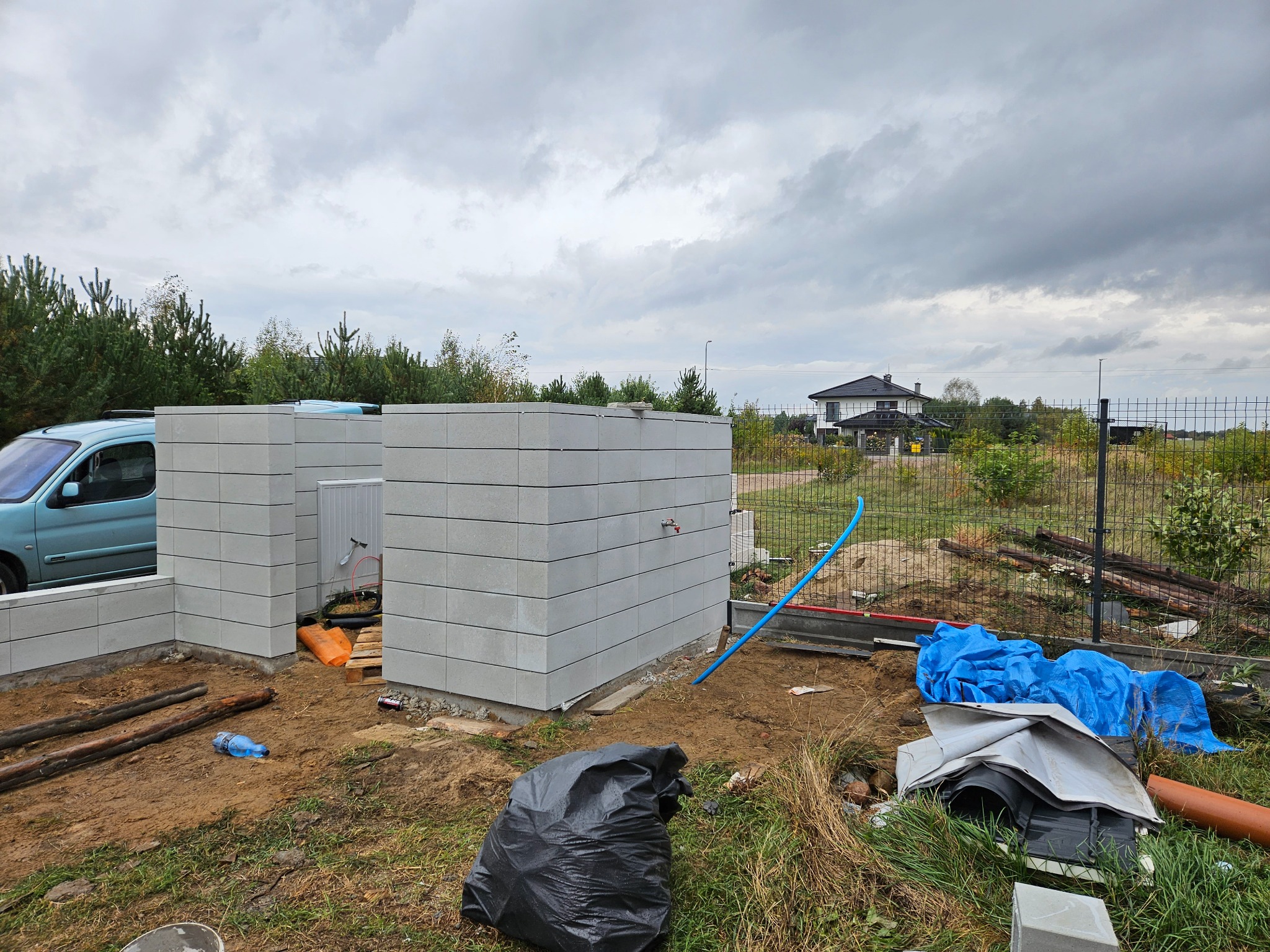 Wznoszenie ścian z szarych bloczków betonowych na fundamencie, widoczne elementy instalacji, teren budowy z gruzem i narzędziami, w tle dom jednorodzinny i pochmurne niebo.