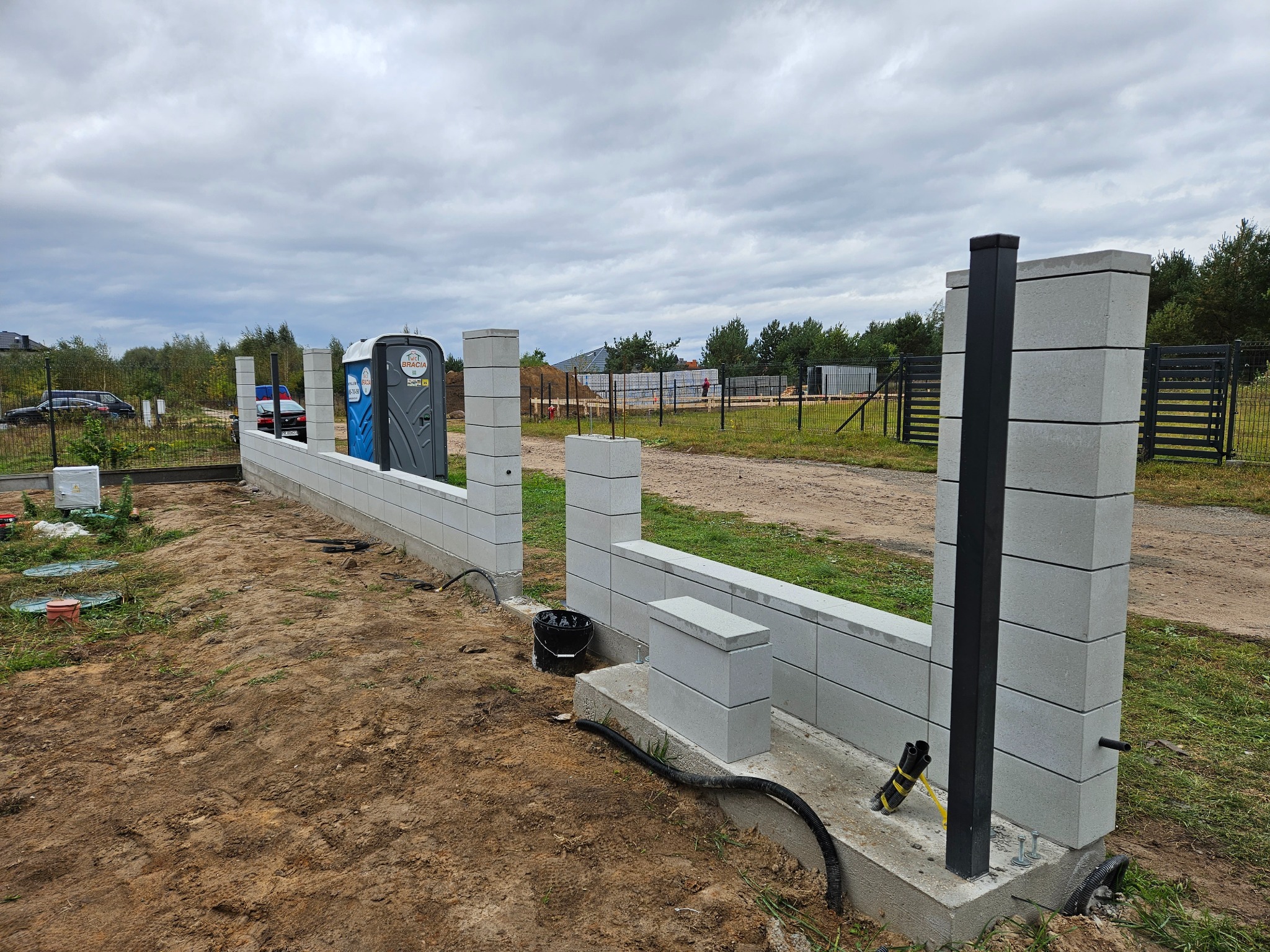 Wznoszenie ogrodzenia z szarej bloczków betonowych, widoczne słupki z czarnymi profilami. W tle las i błękitna toaleta przenośna. Etap budowy.