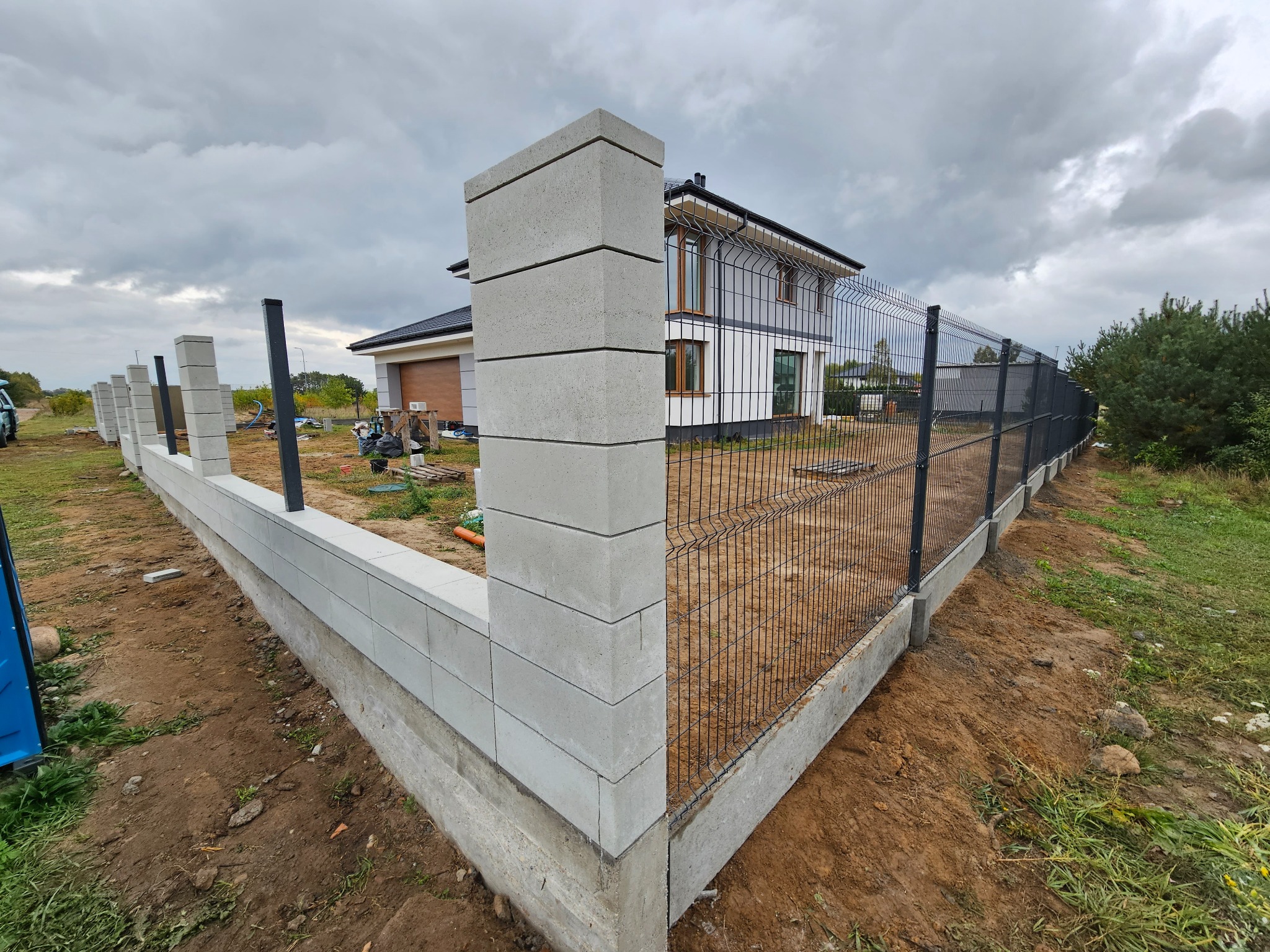 Nowoczesne ogrodzenie panelowe z betonowymi słupkami i podmurówką, otaczające posesję z domem w tle. Widok z perspektywy narożnika ogrodzenia.