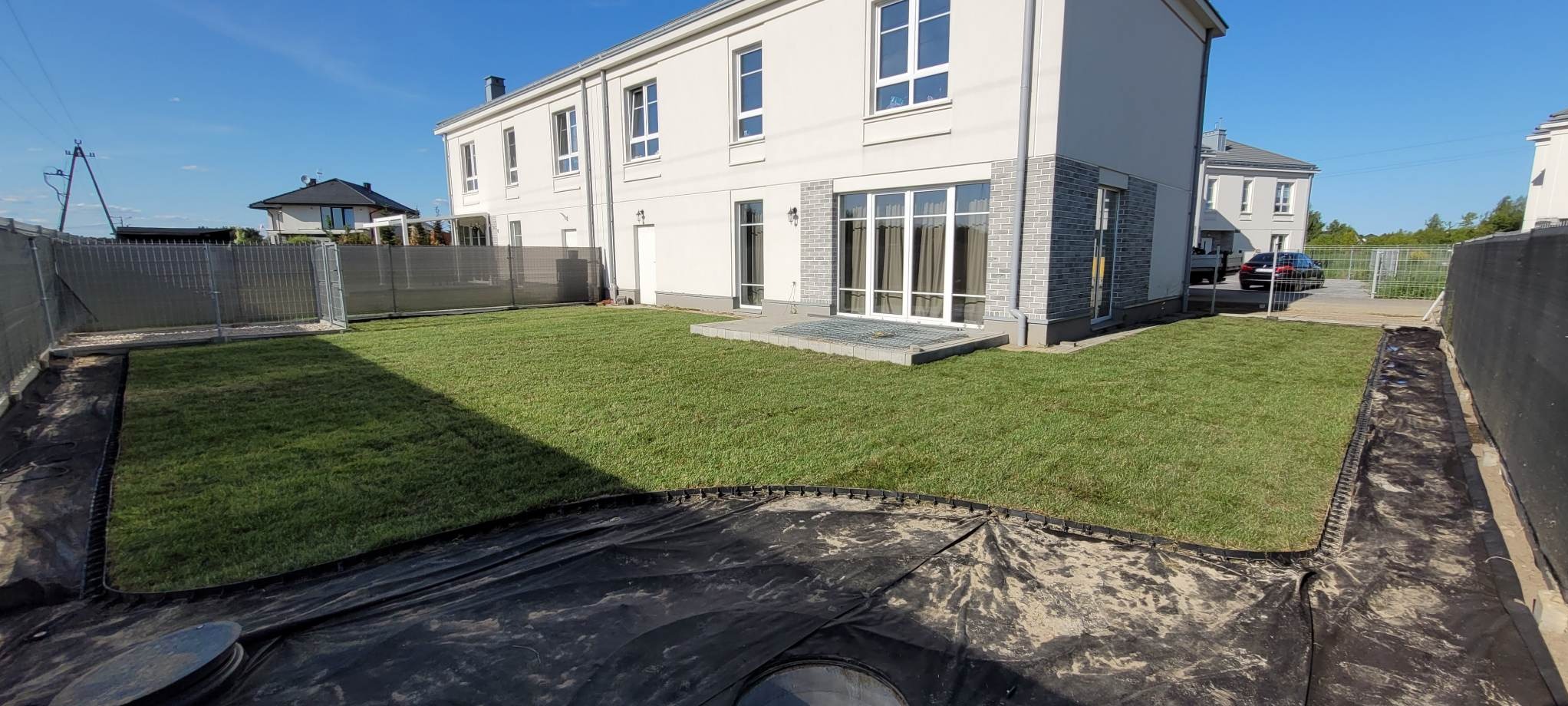 Świeżo ułożony trawnik z rolki przed nowoczesnym domem. Widoczny fragment przygotowanego terenu pod dalsze prace, ogrodzenie w tle i błękitne niebo.