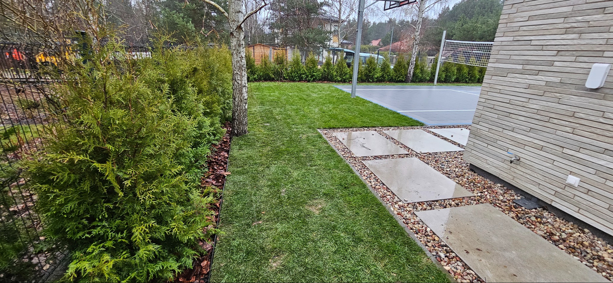 Nowoczesny ogród z równo przystrzyżonym trawnikiem, ścieżką z płyt betonowych i kamieni, oraz żywopłotem z tuj. W tle boisko do siatkówki i budynek z elewacją.