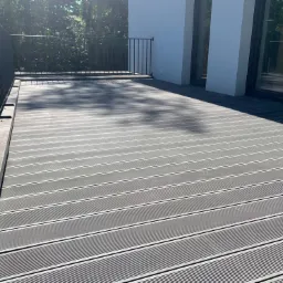 Nowoczesny taras z szarych desek kompozytowych, z czarną metalową balustradą i białymi filarami budynku w tle, oświetlony promieniami słonecznymi tworzącymi cienie.