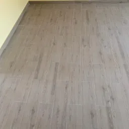 Ułożone, podłużne płytki podłogowe imitujące drewno, z szarą fugą, przy żółtej ścianie z szarą listwą przypodłogową.