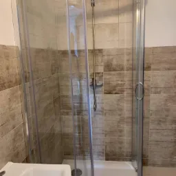 Nowoczesna kabina prysznicowa z przesuwnymi drzwiami, wykończona płytkami w odcieniach beżu i brązu, z widoczną białą umywalką po lewej stronie.