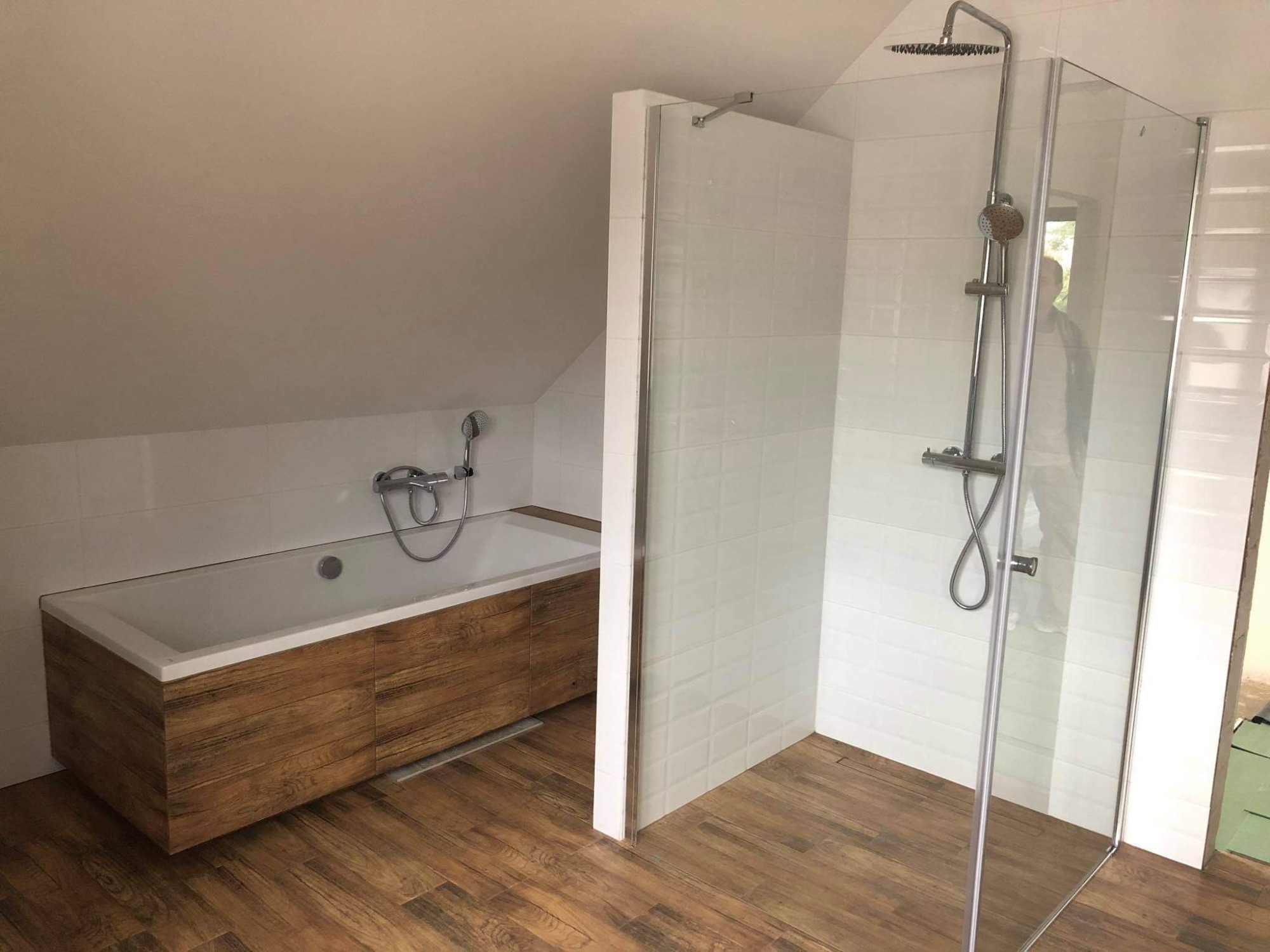 Nowoczesna łazienka na poddaszu z drewnopodobną podłogą, białą wanną obudowaną drewnem i przeszkloną kabiną prysznicową.