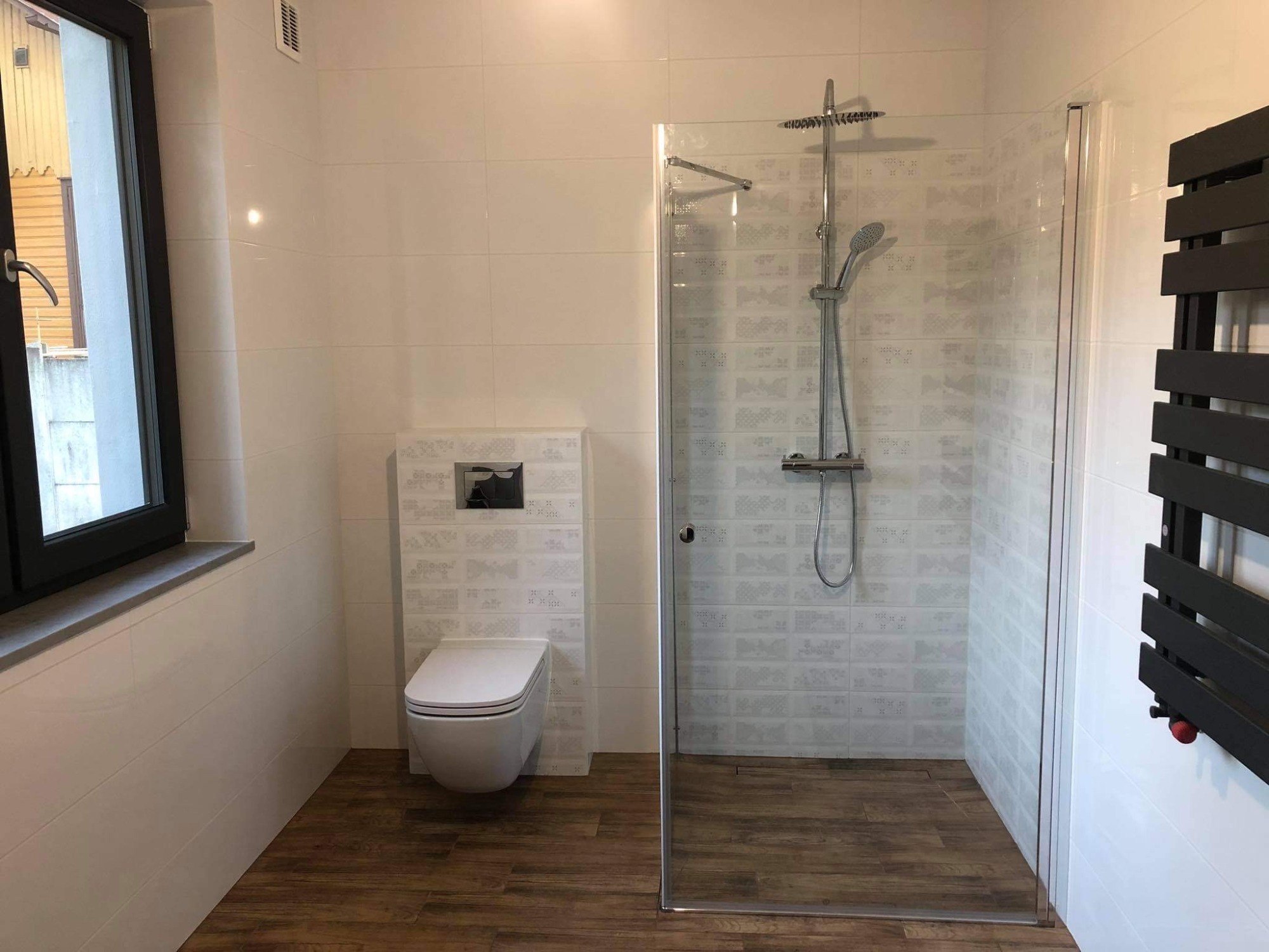 Nowoczesna łazienka z kabiną prysznicową typu walk-in, wiszącą toaletą i grzejnikiem drabinkowym w kolorze antracytowym, wykończona białymi płytkami z delikatnym wzorem i drewnopodobną podłogą.