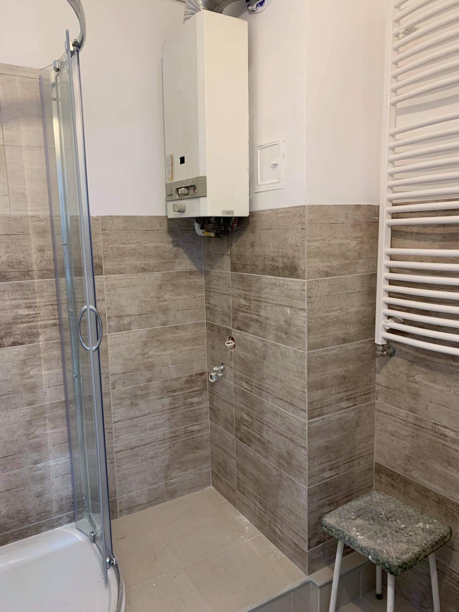 Ukończona łazienka z płytkami imitującymi drewno na ścianach, szklana kabina prysznicowa i biały grzejnik drabinkowy oraz wiszący piec gazowy.