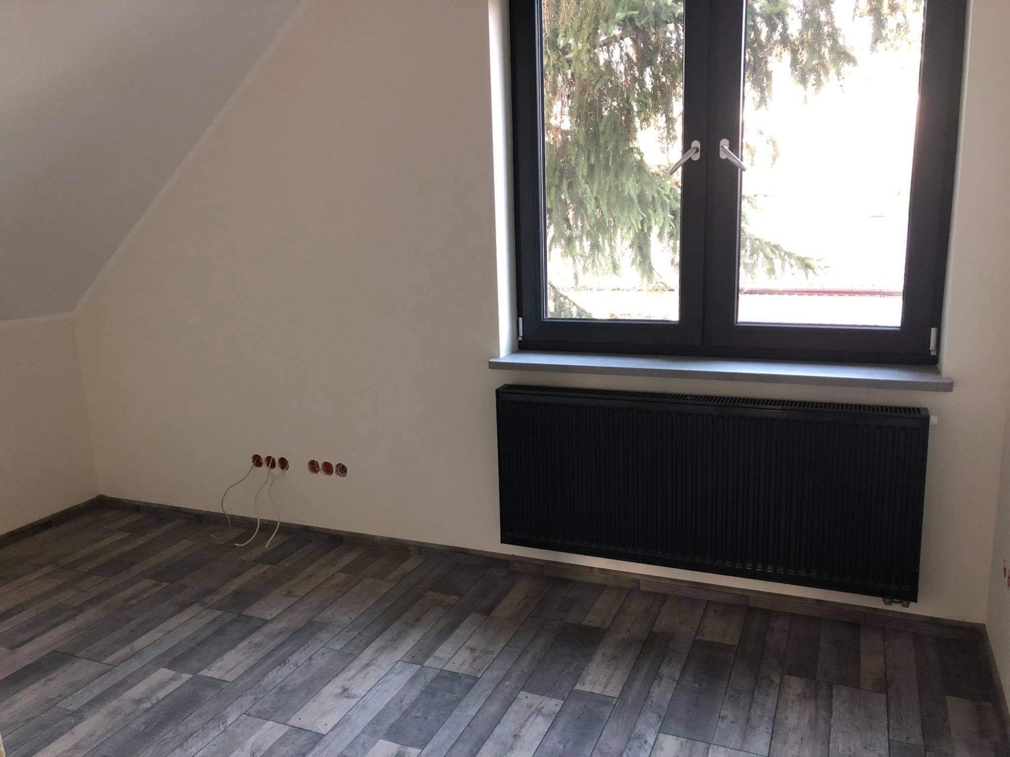 Pokój w trakcie wykańczania: położona podłoga z paneli imitujących drewno, zamontowany czarny grzejnik pod oknem, przygotowane otwory elektryczne w ścianie.