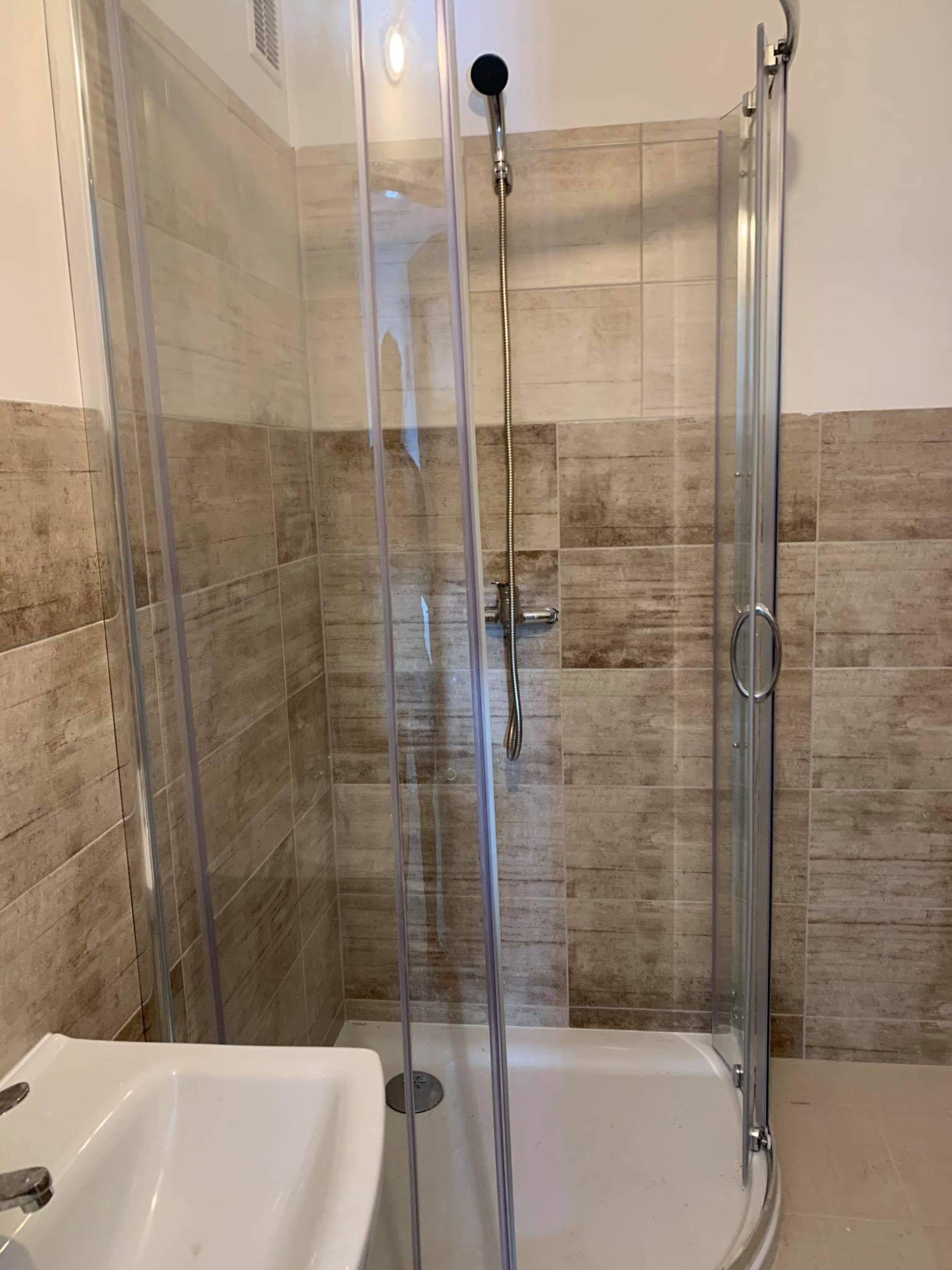 Nowoczesna kabina prysznicowa z przesuwnymi drzwiami, wykończona płytkami w odcieniach beżu i brązu, z widoczną białą umywalką po lewej stronie.