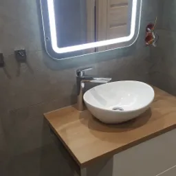 Nowoczesna łazienka z umywalką nablatową, podświetlanym lustrem i szarą okładziną ścienną; widoczne niedokończone podłączenie elektryczne i wiadro z zaprawą w tle.