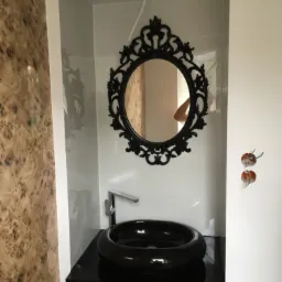 Czarna, okrągła umywalka nablatowa i ozdobne lustro w czarnej ramie w niszy wykończonej płytkami, z widocznymi wystającymi przewodami elektrycznymi.