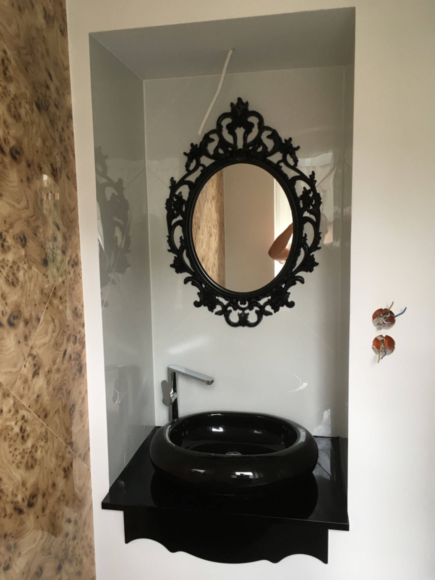 Czarna, okrągła umywalka nablatowa i ozdobne lustro w czarnej ramie w niszy wykończonej płytkami, z widocznymi wystającymi przewodami elektrycznymi.