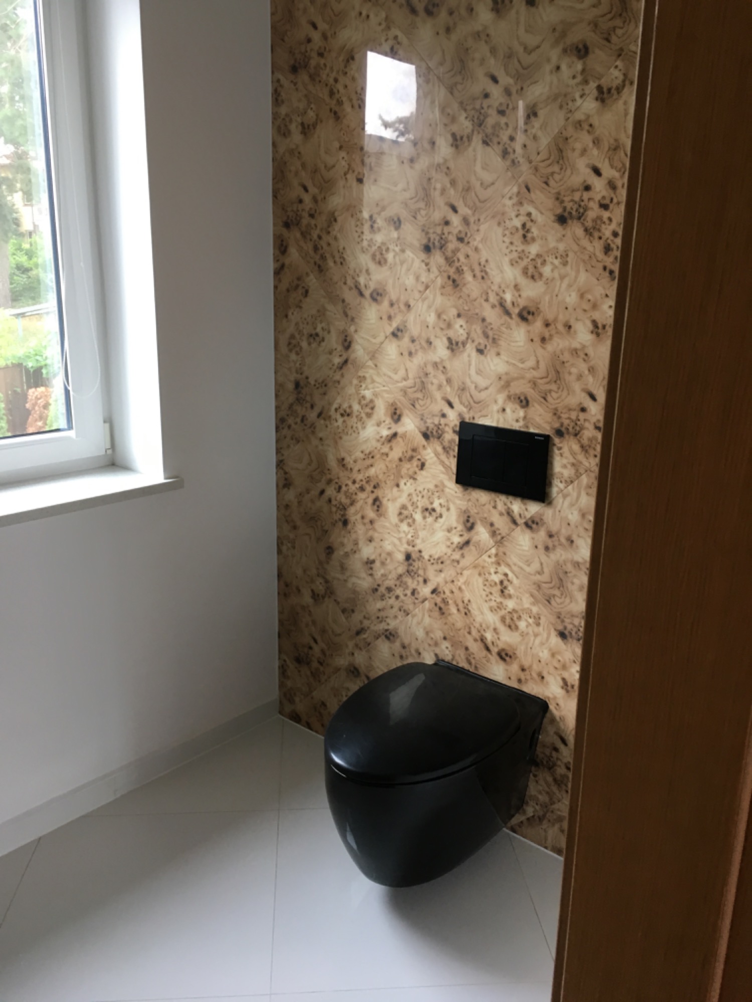 Nowoczesna toaleta w kolorze czarnym, wisząca na ścianie z płytkami imitującymi drewno, biała podłoga i okno z widokiem na zieleń.