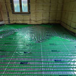 Układanie zielonych rur systemu grzewczego na folii z nadrukiem w surowym wnętrzu budynku z oknem na zewnątrz.