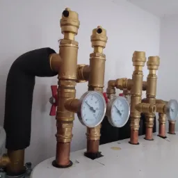 Rozbudowana instalacja hydrauliczna z miedzianymi rurami, zaworami i manometrami, widoczna izolacja termiczna na rurze po lewej stronie.