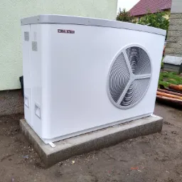 Biała pompa ciepła Stiebel Eltron na betonowym fundamencie, widoczne ślady deszczu na obudowie i błoto wokół, w tle ściana budynku i drewno opałowe.