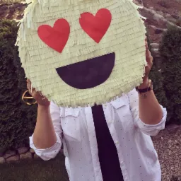 Osoba trzymająca przed twarzą okrągłą piniatę z papierowymi frędzlami, ozdobioną czerwonymi sercami zamiast oczu i czarnym półksiężycem jako uśmiech, na tle zielonych krzewów i kamiennego murku.