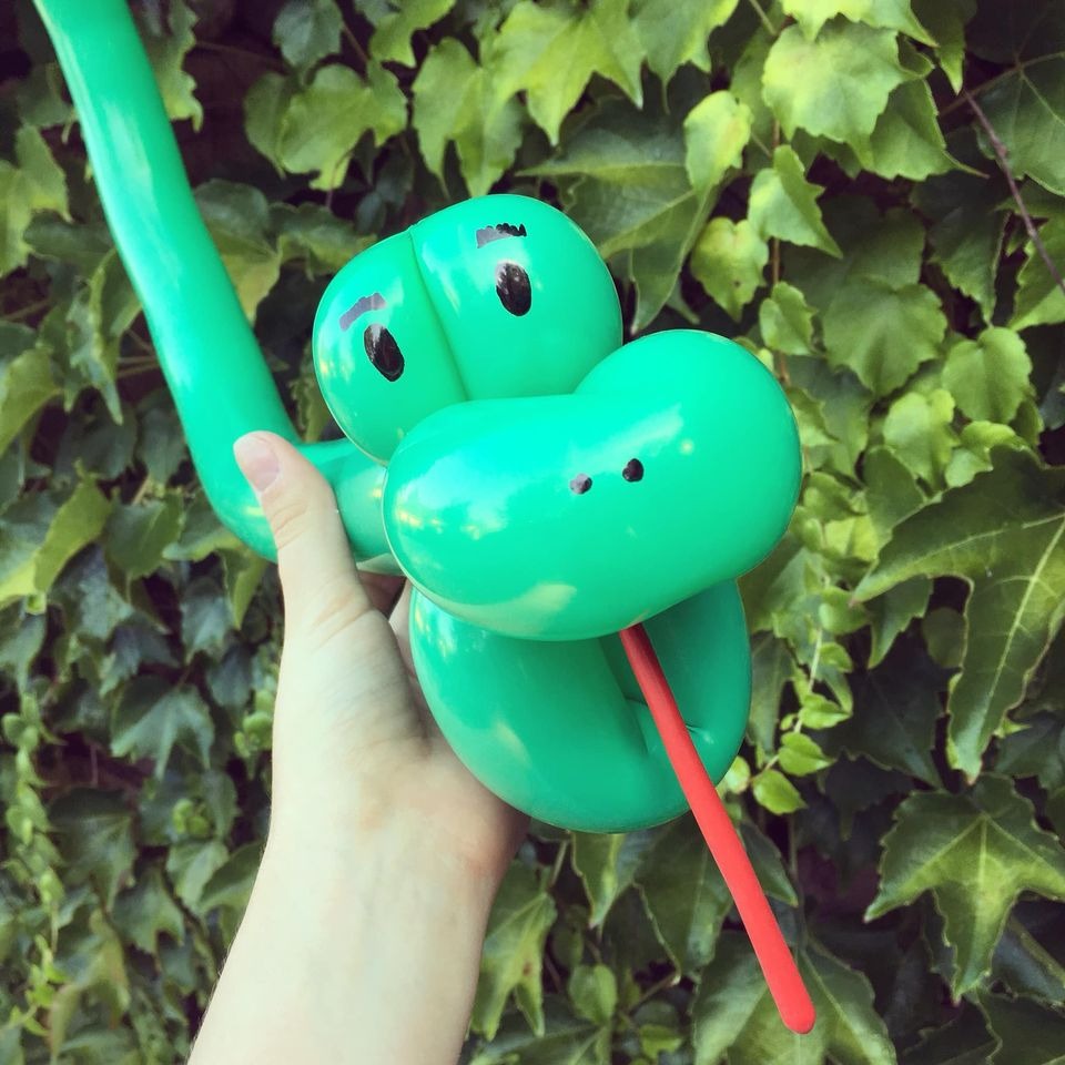 Zielony balonowy krokodyl z czerwonym językiem, trzymany w dłoni na tle ściany z bluszczu.