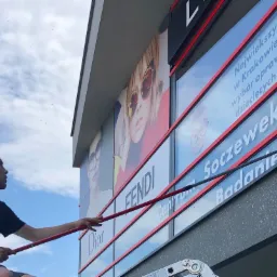 Pracownik myje okna na wysokości w budynku z reklamami Dior i Fendi, używając teleskopowego kija z czerwoną rączką i czarną końcówką, widoczny fragment drabiny.