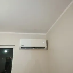 Biała klimatyzacja Kaisai z wyświetlaczem temperatury 30 stopni Celsjusza, zamontowana na beżowej ścianie w rogu pokoju, widoczna ramka okna po lewej stronie.