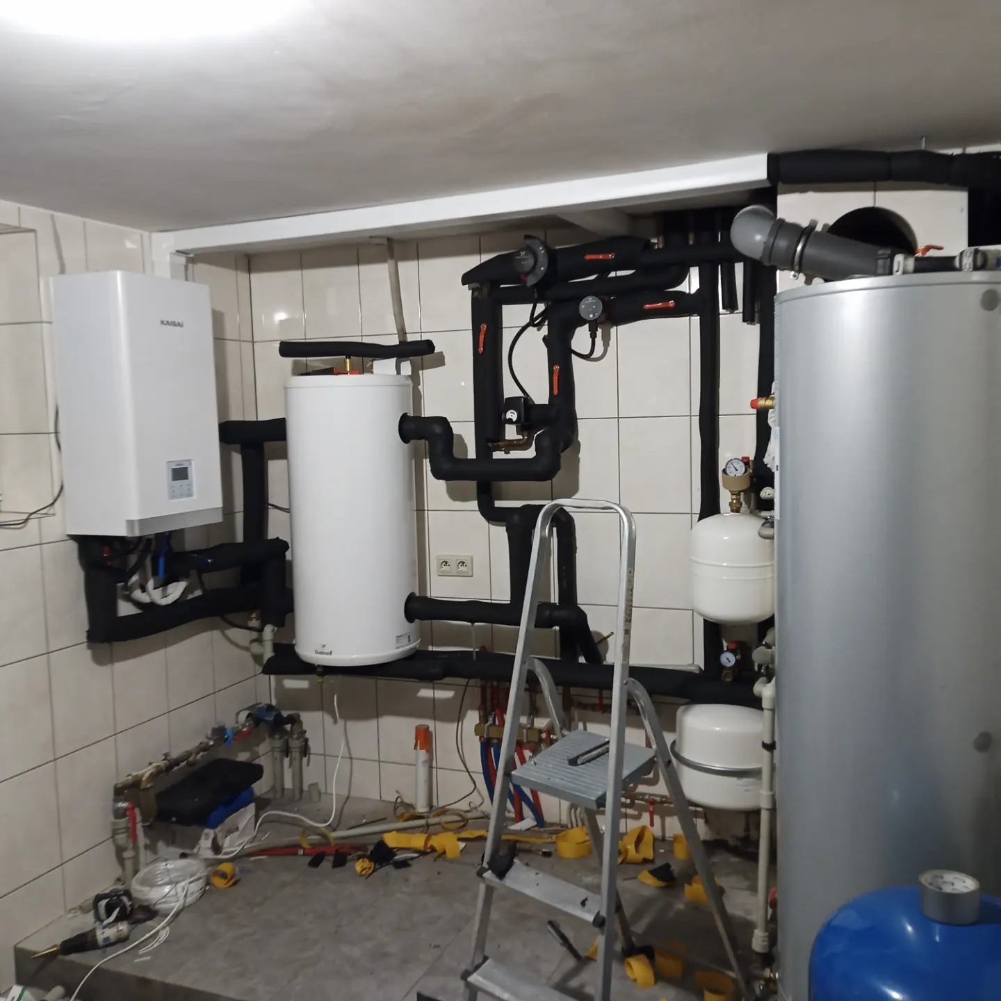 Kotłownia w trakcie montażu: biały piec gazowy Kaisai, biały zasobnik wody Solared na czarnych rurach, srebrny zbiornik buforowy, niebieski zbiornik przeponowy, aluminiowa drabina na tle białych...