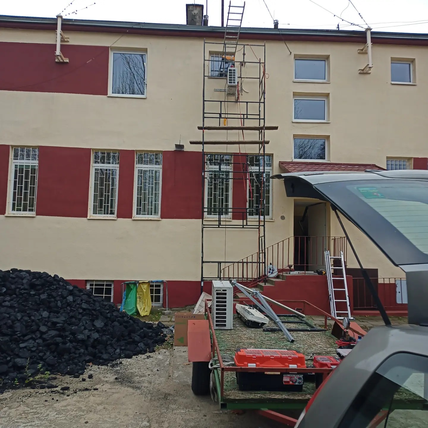Montaż klimatyzatora zewnętrznego na rusztowaniu przy dwukolorowym budynku, widok zza otwartego bagażnika samochodu, w tle hałda węgla i przyczepa z narzędziami.