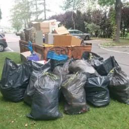 Przepełniony kontener na odpady z gruzem, meblami i kartonami, otoczony stertą czarnych worków na trawniku, widok z boku na tle parkingu i drzew.