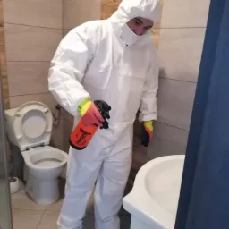 Pracownik w białym kombinezonie ochronnym, masce i rękawicach, trzymający pomarańczowy spryskiwacz w łazience z otwartą toaletą i umywalką.