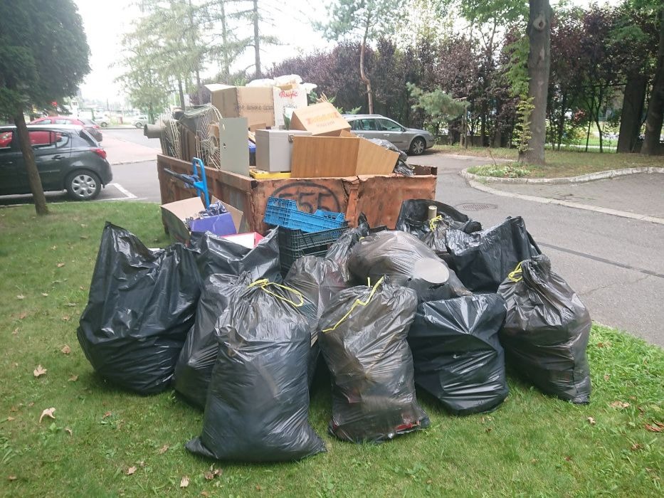 Przepełniony kontener na odpady z gruzem, meblami i kartonami, otoczony stertą czarnych worków na trawniku, widok z boku na tle parkingu i drzew.