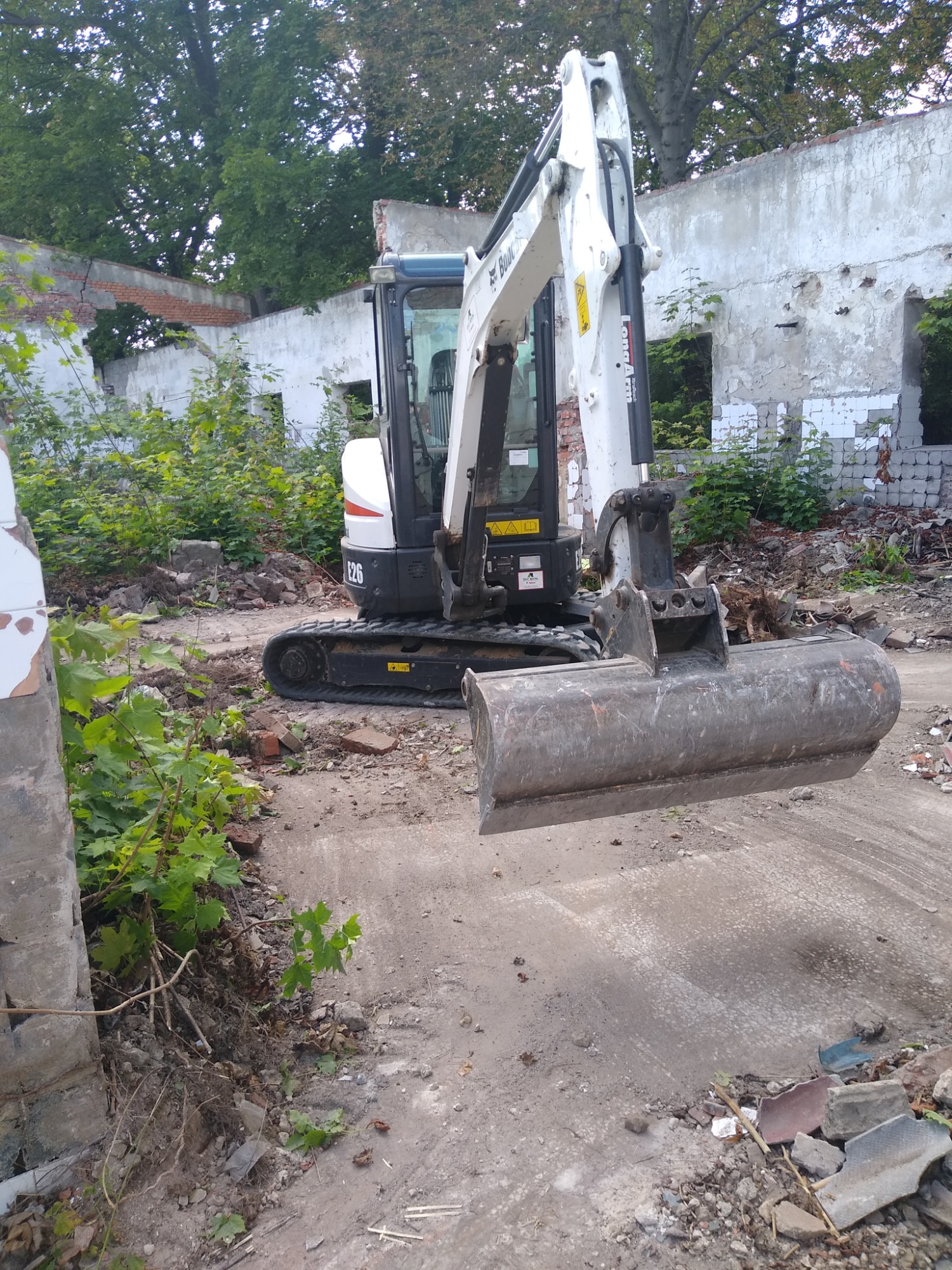 Mała koparka Bobcat E26 na placu budowy z gruzem i fragmentami ścian, otoczona zielenią, przygotowana do dalszych prac rozbiórkowych.