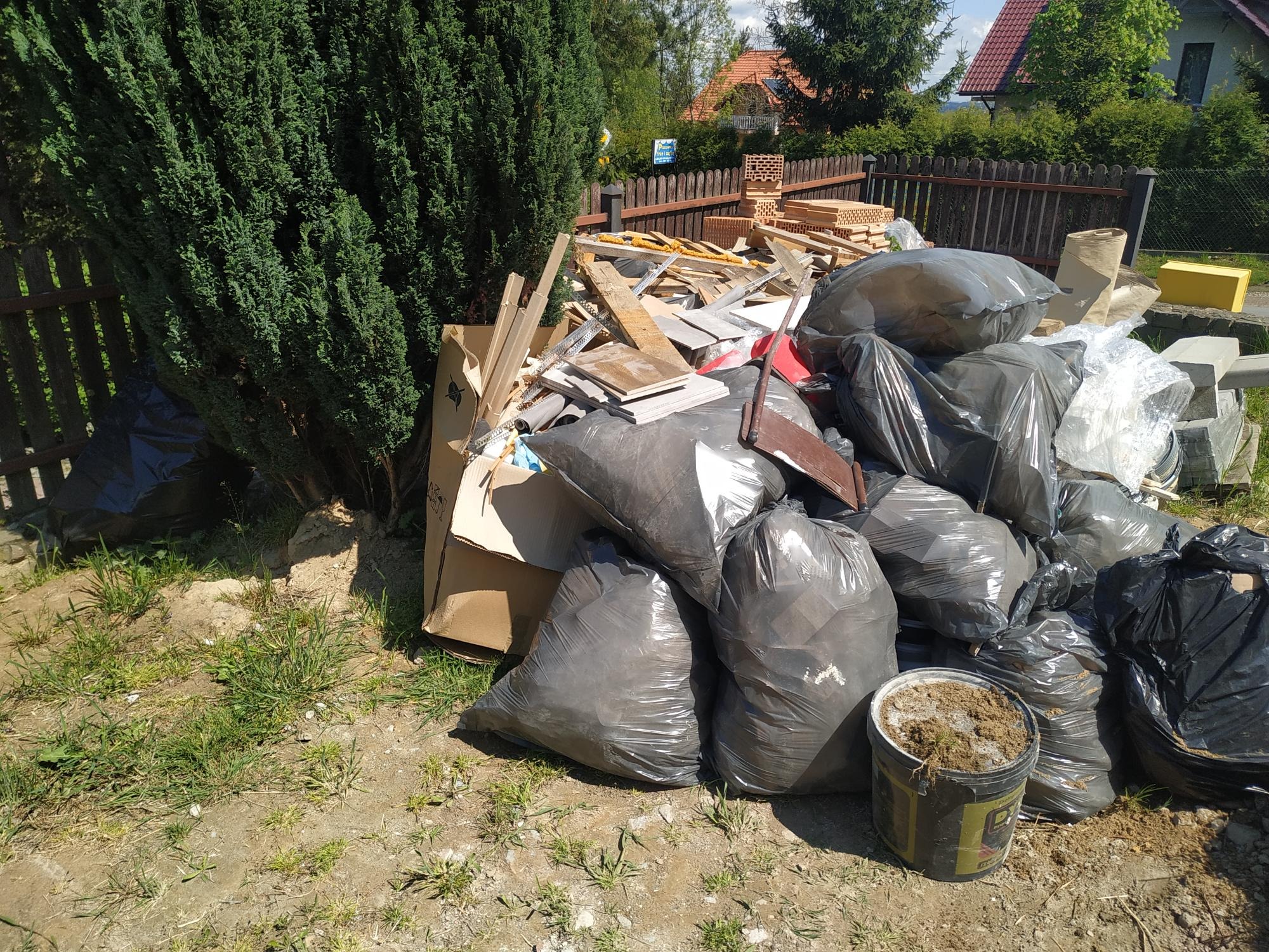 Sterta odpadów budowlanych, w tym cegły, drewno i gruz w czarnych workach foliowych, składowana na trawniku przed ogrodzeniem posesji z widocznym domem w tle.
