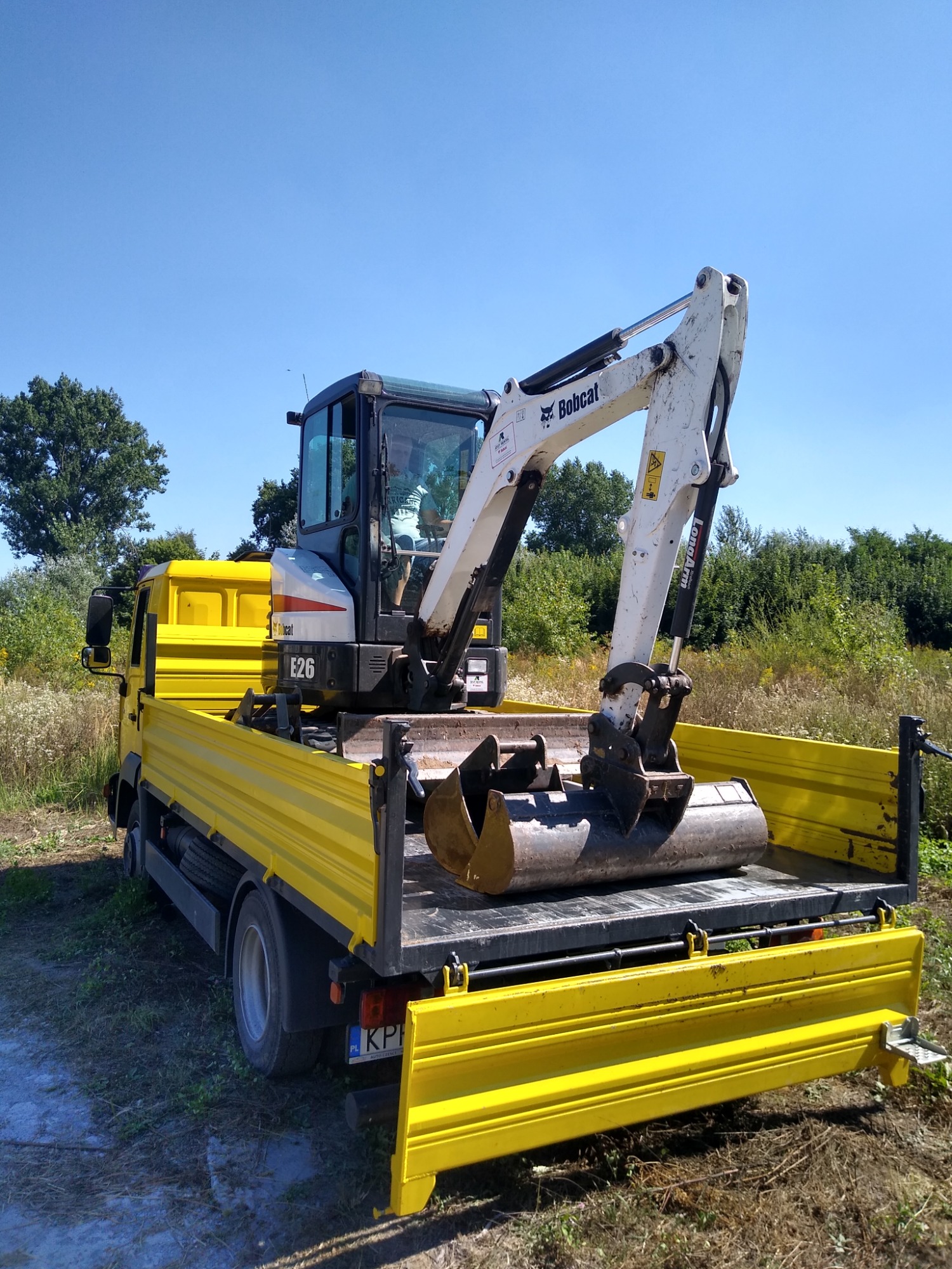 Żółta ciężarówka z otwartą skrzynią ładunkową, na której stoi minikoparka Bobcat E26, widok z tyłu, błękitne niebo w tle.