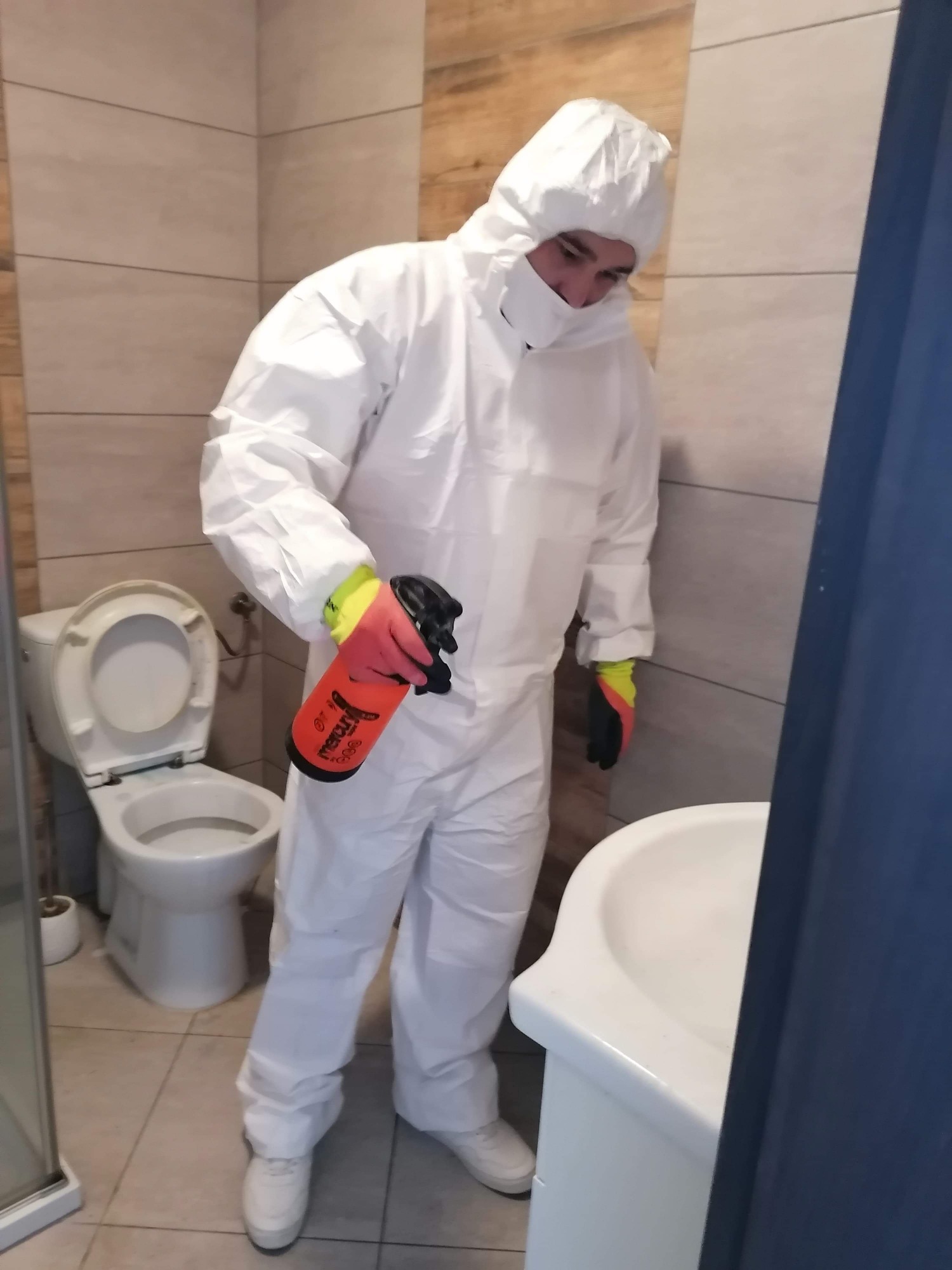 Pracownik w białym kombinezonie ochronnym, masce i rękawicach, trzymający pomarańczowy spryskiwacz w łazience z otwartą toaletą i umywalką.