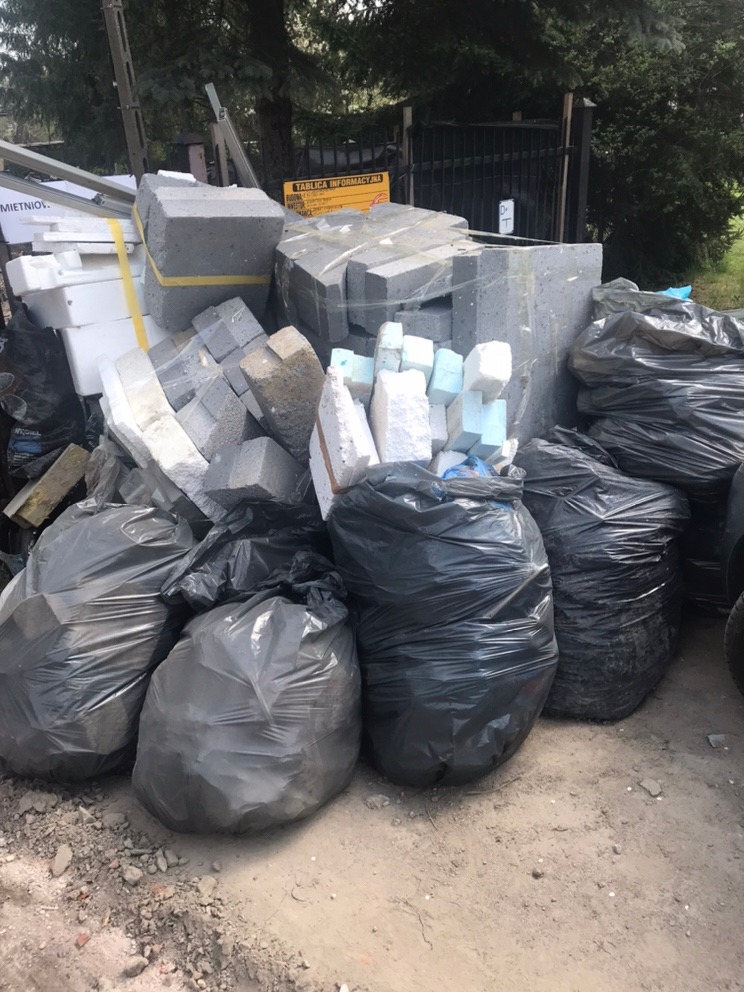 Sterta gruzu budowlanego i styropianu w czarnych workach oraz luzem, częściowo zabezpieczona żółtą taśmą, złożona na ziemi obok metalowego ogrodzenia.