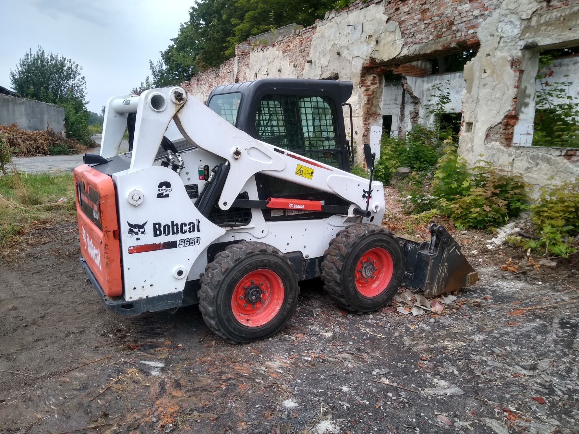 Kompaktowa ładowarka Bobcat S650 z czerwonymi felgami stoi na placu budowy z gruzem i fragmentami cegieł, w tle zrujnowany budynek bez dachu i okien.