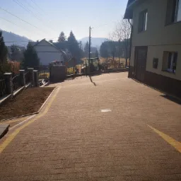 Nowo ułożona, brązowa kostka brukowa z żółtymi pasami wyznaczającymi kierunek, prowadząca do domu. W tle widoczne maszyny budowlane, ogrodzenie i krajobraz górski.