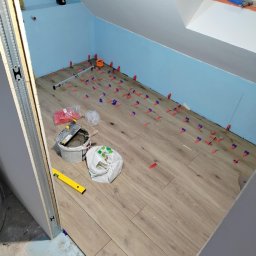 Usługi ogólno-budowlane - Układanie paneli podłogowych w pokoju na poddaszu. Widoczne narzędzia, poziomica i niebieskie klipsy dystansowe. Ściany pomalowane na jasny niebieski kolor.