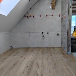 Usługi ogólno-budowlane - Wykończenie łazienki na poddaszu: ułożone płytki imitujące marmur z poziomowaniem, drewnopodobna podłoga, okno dachowe. Widoczne elementy instalacji.