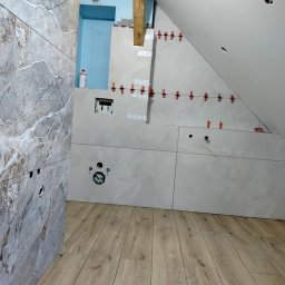 Usługi ogólno-budowlane - Układanie płytek w łazience na poddaszu. Widoczne płytki imitujące marmur na ścianie i podłodze, oraz białe płytki na skośnej ścianie z czerwonymi krzyżykami dystansowymi.