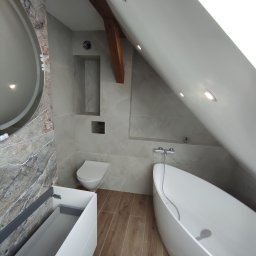 Usługi ogólno-budowlane - Nowoczesna łazienka na poddaszu z wolnostojącą wanną, marmurową ścianą z okrągłym lustrem i drewnianą belką stropową. Umywalka z szafką i toaleta w neutralnej kolorystyce.