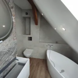 Nowoczesna łazienka na poddaszu z wolnostojącą wanną, marmurową ścianą z okrągłym lustrem i drewnianą belką stropową. Umywalka z szafką i toaleta w neutralnej kolorystyce.