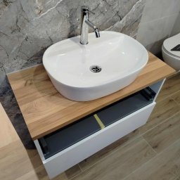 Usługi ogólno-budowlane - Nowoczesna łazienka: biała, owalna umywalka nablatowa z chromowaną baterią na drewnianym blacie, szara marmurowa ściana i drewnopodobna podłoga.