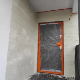 Elewacja z fakturą drewna, zabezpieczone okno folią malarską. Detal wykończenia ściany zewnętrznej z widoczną strukturą desek. Prace malarskie w toku.