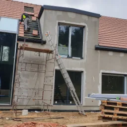 Montaż brązowej dachówki na nowym domu z dużym oknem, widoczne rusztowania i drabina oparta o elewację w trakcie tynkowania.