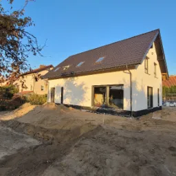 Nowoczesny dom jednorodzinny z jasną elewacją i brązowym dachem, widoczny wczesną jesienią na tle błękitnego nieba, z przygotowanym terenem wokół budynku.